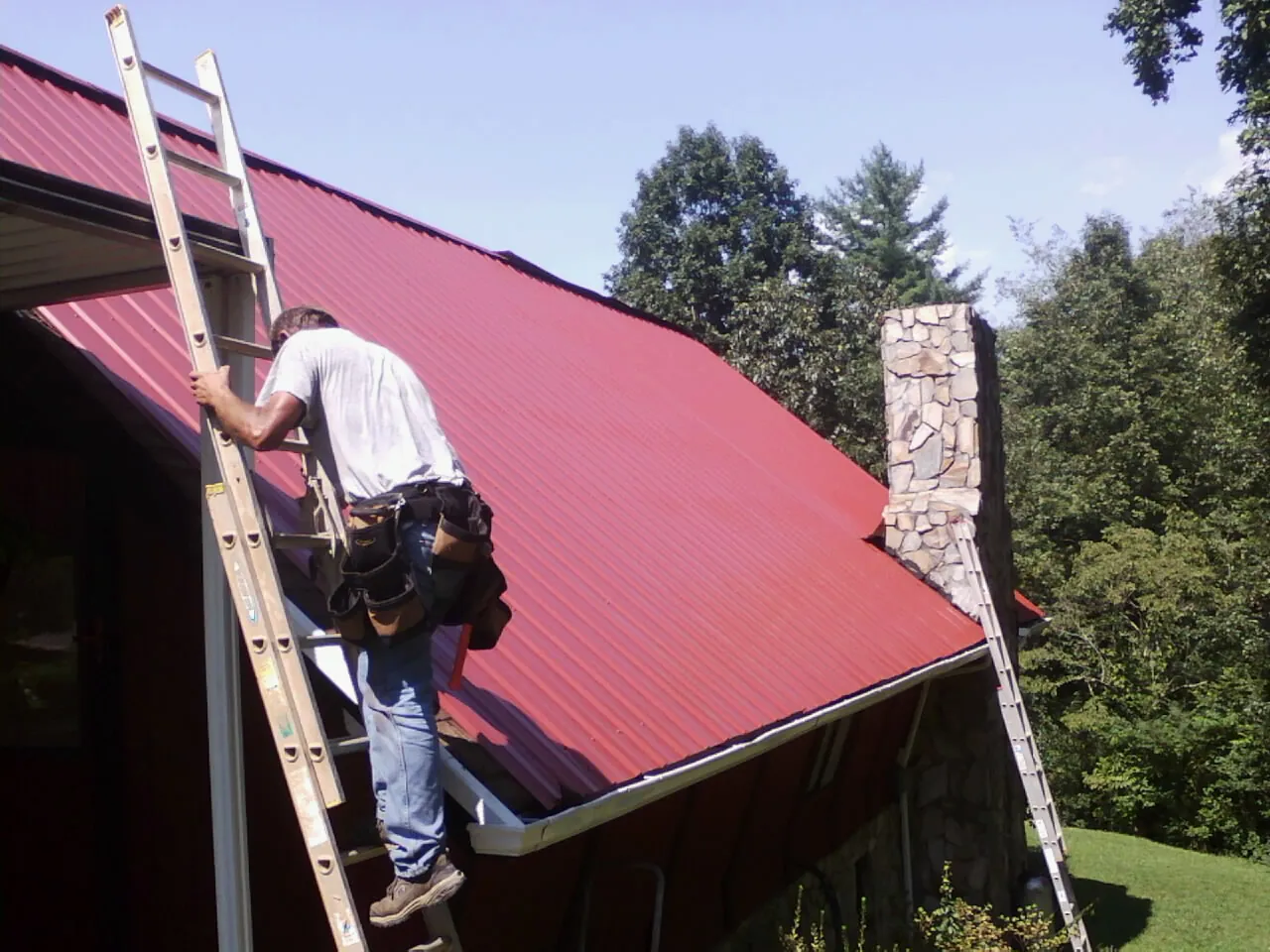 Roofer on a ladder beside a red metal roof and stone chimney — Soffit & Fascia Repair by Roofing Brothers in East Rutherford